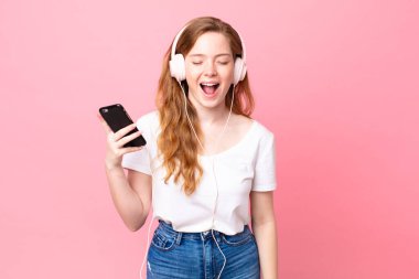 Güzel kızıl saçlı kadın agresif bir şekilde bağırıyor, kulaklıkları ve akıllı telefonlarıyla çok kızgın görünüyor.
