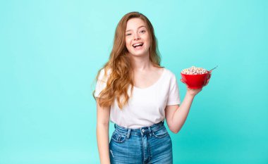 Güzel kızıl saçlı kadın mutlu görünüyor ve hoş bir şekilde şaşırmış ve elinde bir kahvaltı kasesi tutuyor.