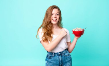 Güzel kızıl saçlı kadın kendini mutlu hissediyor ve meydan okumayla yüzleşiyor ya da kutlama yapıyor ve elinde bir kahvaltı kasesi tutuyor.