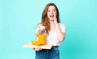 Güzel kızıl saçlı kadın mutlu, elleri ağzının yanında ve elinde portakal suyu tutarak bağırıyor.