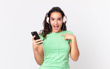 Güzel İspanyol bir kadın kendini mutlu hissediyor ve kulaklıklar ve akıllı telefonlarla kendini işaret ediyor.