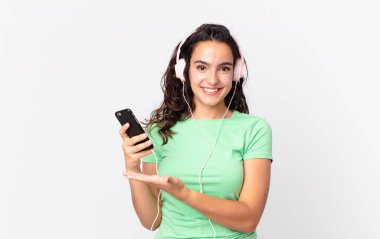 Güzel İspanyol bir kadın neşeli bir şekilde gülümsüyor, mutlu hissediyor ve kulaklık ve akıllı telefonla bir konsept gösteriyor.