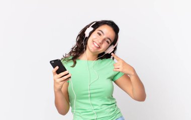 Güzel İspanyol bir kadın, kendine güvenerek gülümsüyor. Kendi gülüşünü işaret ediyor. Kulaklığı ve akıllı telefonu var.