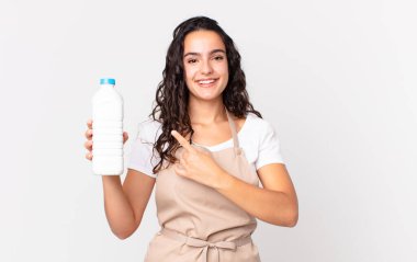 İspanyol güzel aşçı kadın neşeyle gülümsüyor, mutlu hissediyor ve bir tarafı işaret ediyor ve elinde bir süt şişesi tutuyor.