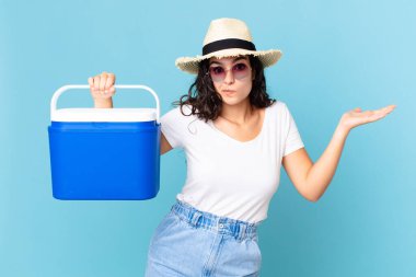 Güzel İspanyol kadın şaşkın, kafası karışmış ve elinde taşınabilir bir buzdolabı tutmaktan şüphe duyuyor.