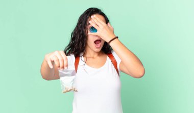 İspanyol görünümlü güzel bir kadın şok olmuş, korkmuş ya da dehşete düşmüş gibi görünüyor, yüzünü kaplamış ve elinde bir marihuana çantası tutuyor.