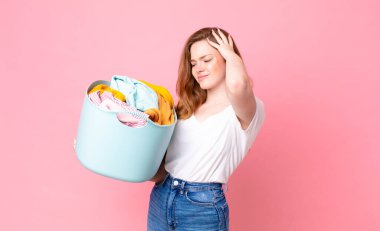 Kızıl saçlı güzel kadın şaşkın ve kafası karışmış hissediyor, kafasını kaşıyor ve çamaşır sepetini kıyafetlerle tutuyor.