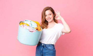 Kızıl saçlı, güzel bir kadın mutlu. Onayını gösteriyor. İyi bir jest yapıyor ve çamaşır sepetini kıyafetlerle tutuyor.