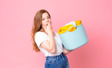 Kızıl saçlı, güzel bir kadın elleri ve elleri ile ağzını kapatıyor ve çamaşır sepetini kıyafetlerle tutuyor.