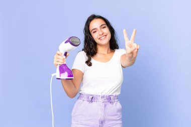 İspanyol güzel kadın gülümsüyor ve arkadaş canlısı görünüyor. İki numarayı gösteriyor ve elinde demir buhar tutuyor.