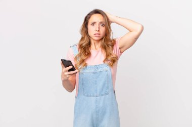 Güzel bir kadın kendini stresli, endişeli ya da korkmuş hissediyor. Elleri başının üstünde ve elinde akıllı bir telefon tutuyor.