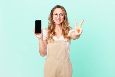Güzel bir kadın gülümsüyor ve mutlu görünüyor, zafer ya da barış işareti yapıyor ve akıllı bir telefon tutuyor.
