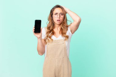 Güzel bir kadın kendini stresli, endişeli ya da korkmuş hissediyor. Elleri başının üstünde ve elinde akıllı bir telefon tutuyor.