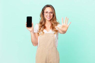 Güzel bir kadın gülümsüyor ve arkadaş canlısı görünüyor, 5 numarayı gösteriyor ve elinde bir akıllı telefon var.