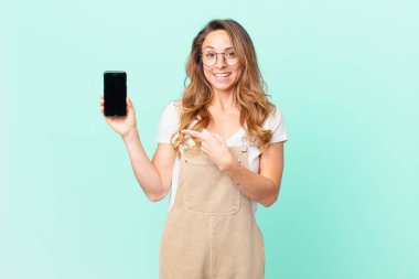 Güzel bir kadın heyecanlı ve şaşırmış görünüyor. Yan tarafı gösteriyor ve elinde bir akıllı telefon tutuyor.