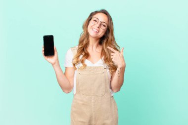 Güzel bir kadın kendine güvenerek gülümsüyor. Kendi gülüşünü ve akıllı telefonunu gösteriyor.