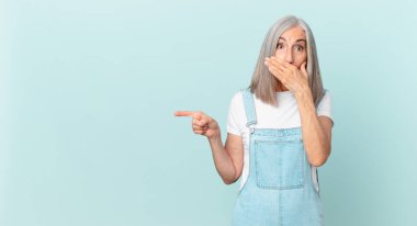 Orta yaşlı, beyaz saçlı bir kadın elleri ile ağzını kapatıyor ve bir yandan işaret ediyor.