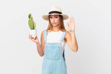 Güzel bir kadın ciddi görünüyor. Avuç içi gösteriyor. Jesti durduruyor ve elinde kaktüs bitkisi tutuyor.