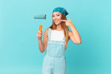 Güzel bir kadın, kendine güvenerek gülümseyerek yeni bir ev duvarı boyayarak kendi geniş gülümsemesine işaret ediyor.