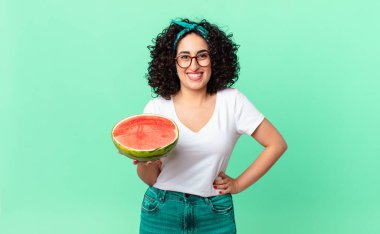 Güzel Arap kadın mutlu bir şekilde gülümsüyor, eli kalçasında, kendinden emin ve elinde bir karpuz tutuyor. yaz konsepti