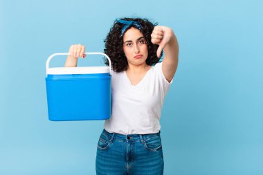 Güzel bir Arap kadın kızgın hissediyor, baş parmaklarını gösteriyor ve portatif bir buzdolabı tutuyor.