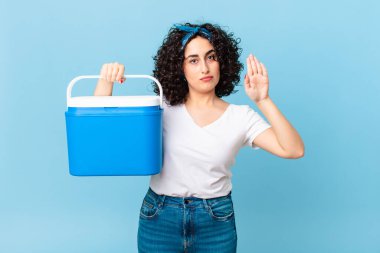 Güzel bir Arap kadın ciddi görünüyor. Avuç içi açık, el hareketi yapıyor ve portatif bir buzdolabı tutuyor.