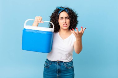Güzel bir Arap kadın kızgın, sinirli ve hayal kırıklığına uğramış görünüyor ve portatif bir buzdolabı tutuyor.