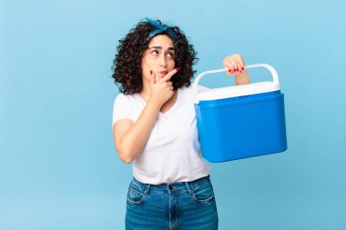 Güzel Arap kadın düşünüyor, kuşkucu ve şaşkın hissediyor ve portatif bir buzdolabı tutuyor.