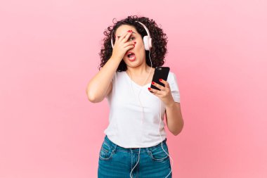 Güzel bir Arap kadını şok olmuş, korkmuş ya da korkmuş görünüyor. Yüzü kulaklıkla ve akıllı telefonla örtülmüş.