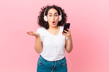 Güzel bir Arap kadın kulaklıklar ve akıllı telefonuyla şok olmuş ve şaşırmış hissediyor.
