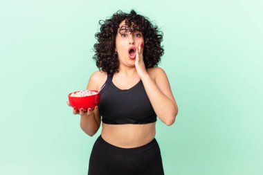 Güzel Arap kadın şok olmuş ve korkmuş. Spor kıyafetleri giyiyor ve elinde mısır gevreği kasesi tutuyor.