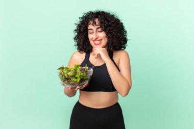 Güzel bir Arap kadın mutlu, kendinden emin bir ifadeyle gülümsüyor. Çenesinde elleri ve elinde salatayla. diyet kavramı