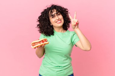 Güzel Arap kadın gülümsüyor ve arkadaş canlısı görünüyor. İki numarayı gösteriyor ve elinde sosisli sandviç tutuyor.
