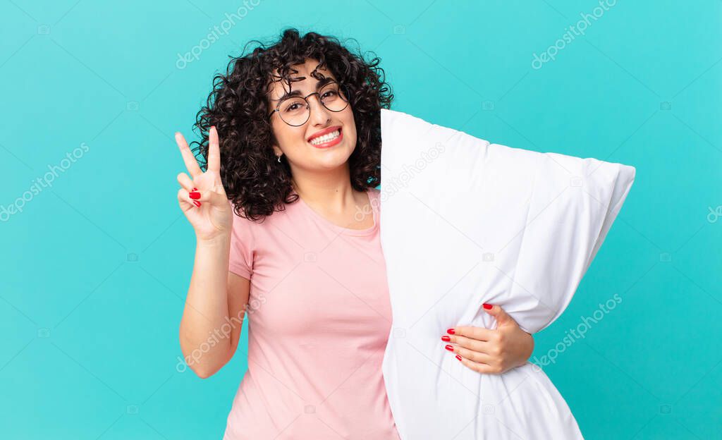 pretty arab woman smiling and looking friendly, showing number two. wearing pajamas and holding a pillow