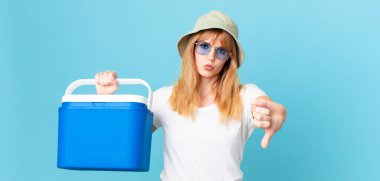Kızıl saçlı güzel bir kadın kendini kötü hissediyor, baş parmağını kaldırıyor ve portatif bir buzdolabı tutuyor. yaz konsepti