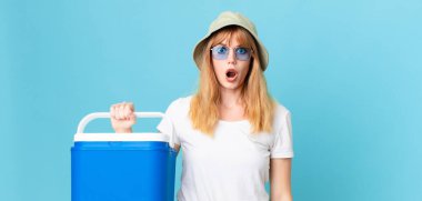 Güzel kızıl saçlı kadın şok olmuş ya da şaşırmış görünüyor ve elinde taşınabilir bir buzdolabı tutuyor. yaz konsepti