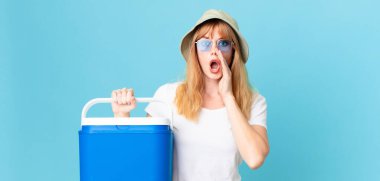 Güzel kızıl saçlı kadın mutlu hissediyor, elleri ağızının yanında ve portatif bir buzdolabı tutarak bağırıyor. yaz konsepti