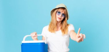 Güzel kızıl saçlı kadın gülümsüyor ve arkadaş canlısı görünüyor. Bir numarayı gösteriyor ve portatif bir buzdolabı tutuyor. yaz konsepti