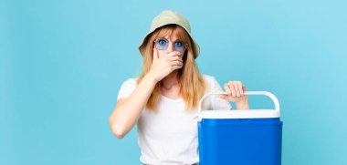 Güzel kızıl saçlı kadın elleri ve elleri ile ağzını kapatıyor ve portatif bir buzdolabı tutuyor. yaz konsepti