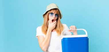 Güzel kızıl saçlı, ağzı ve gözleri açık, elleri çenesinde ve portatif bir buzdolabı tutan bir kadın. yaz konsepti