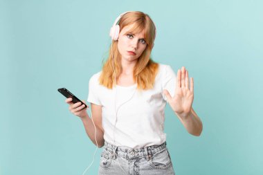 Güzel kızıl saçlı kadın ciddi görünüyor. Avuç içini gösteriyor. Durma hareketi yapıyor ve kulaklıkla müzik dinliyor.