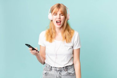 Güzel kızıl saçlı kadın agresif bağırıyor, çok kızgın görünüyor ve kulaklıkla müzik dinliyor.