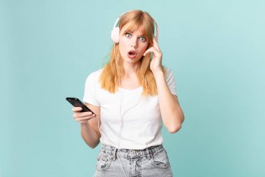 Güzel kızıl saçlı kadın şaşırmış görünüyor, yeni bir düşünce, fikir ya da konsept fark ediyor ve kulaklıkla müzik dinliyor.