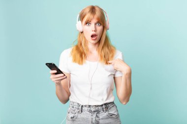 Güzel kızıl saçlı kadın ağzı açık, şok olmuş ve şaşırmış görünüyor, kendini işaret ediyor ve kulaklıkla müzik dinliyor.