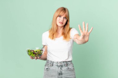Güzel kızıl saçlı kadın gülümsüyor ve arkadaş canlısı görünüyor. Beş numarayı gösteriyor ve elinde salata tutuyor. diyet kavramı