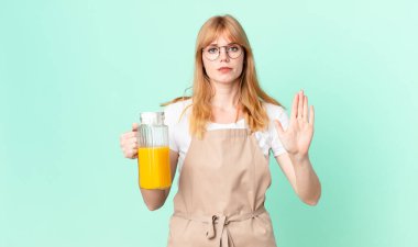 Güzel kızıl saçlı kadın ciddi görünüyor. Avuç içini gösteriyor. Önlük takıp portakal suyu hazırlıyor.