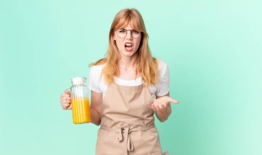 Güzel kızıl saçlı kadın kızgın, sinirli ve sinirli görünüyor. Portakal suyu hazırlayan önlükten.