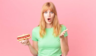 Güzel kızıl saçlı kadın şok olmuş, şaşırmış ve elinde sosisli sandviç tutuyor.