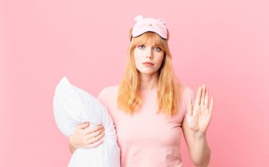 Güzel kızıl saçlı kadın ciddi görünüyor. Avuç içini gösteriyor. Durdurma hareketi yapıyor. Pijama giyiyor ve yastık tutuyor.