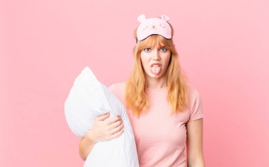 Güzel kızıl saçlı kadın iğreniyor, sinirleniyor ve dili dışarı çıkıyor. Pijama giyiyor ve yastık tutuyor.
